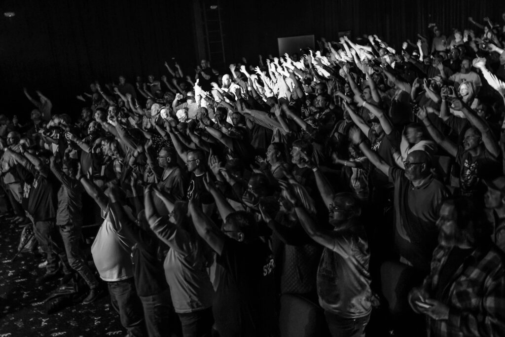 A packed audience with hands raised at a concert in the theater